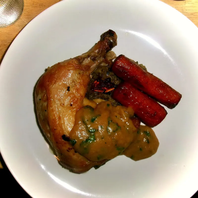 Roast chicken leg under a thick, herb-flecked sauce, with glazed carrots, on a white plate.