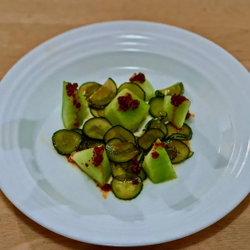 Sliced cucumber and cubes of pale green melon dotted with chili crisp, on a white plate.