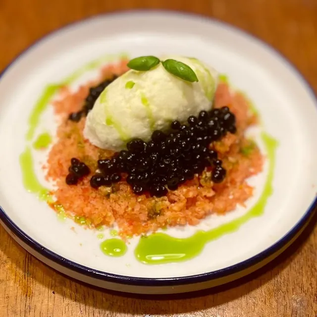 A deconstructed caprese: a scoop of white cream with basil, dark pearls over chopped tomato, and drops of green oil, in a blue-rimmed bowl.