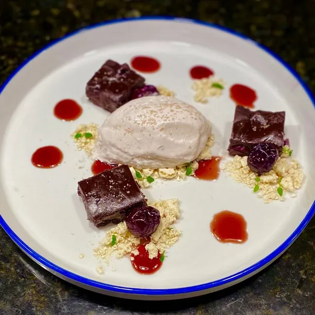 Squares of dark chocolate, a quenelle of pale ice cream, cherry pieces and dots of red sauce over crumbs on a blue-rimmed plate.