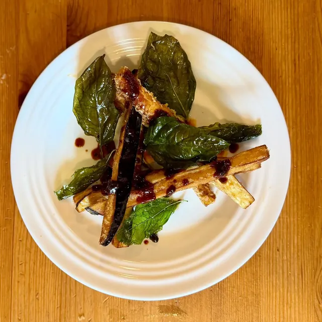 Fried eggplant batons with crisp fried basil leaves and drops of a dark glaze on a ridged white plate.