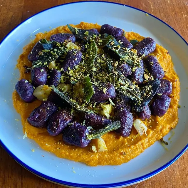 Purple gnocchi with charred greens and herbs over a bright orange carrot purée on a blue-rimmed plate.
