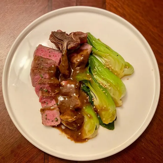 Sliced pink lamb in a dark glaze beside braised bok choy on a white plate.