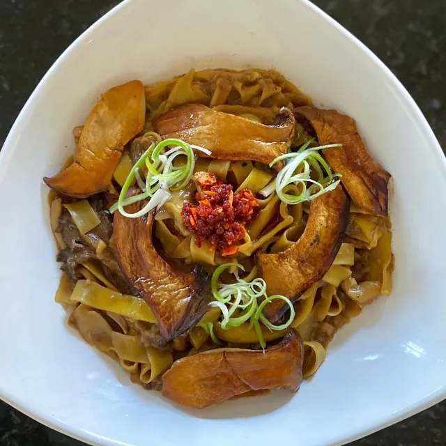 Wide noodles with sautéed mushrooms, scallion curls and chili flakes in a white bowl.