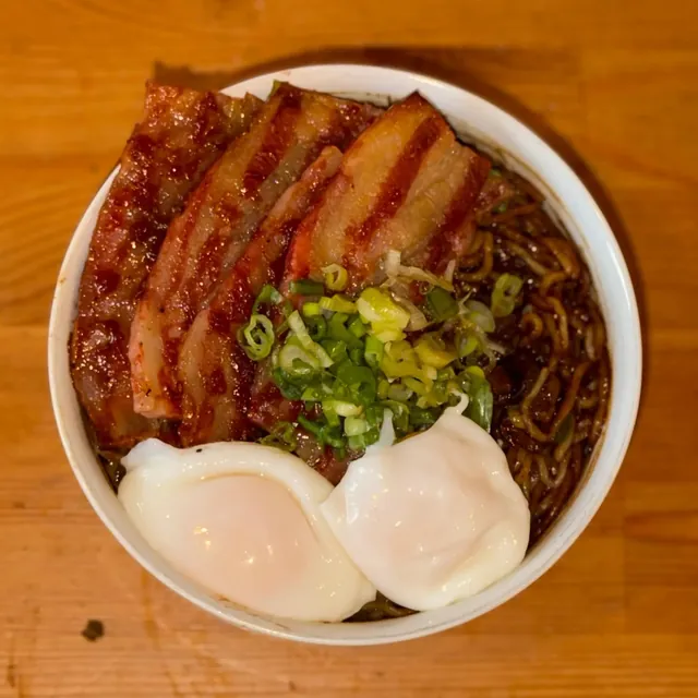 Bowl of dark, soy-glazed noodles topped with slices of grilled pork belly, a heap of scallions, and two poached eggs.