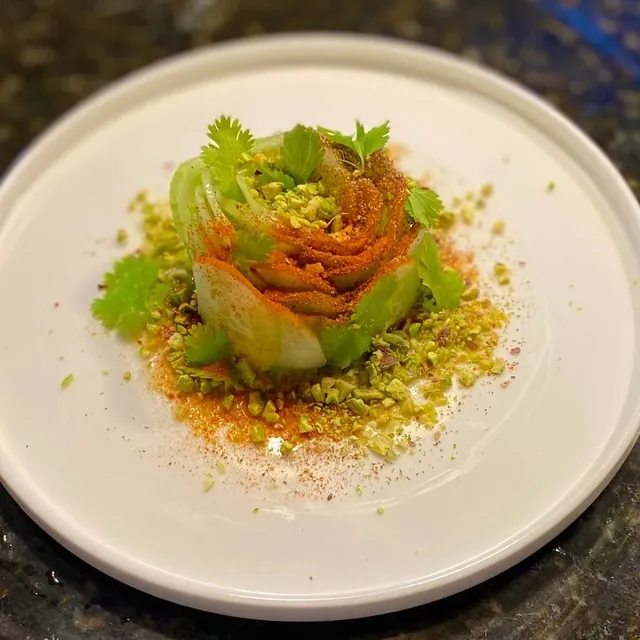 A tall, tightly built salad of greens and herbs with a coral dressing, ringed with crushed pistachio, on a white plate.