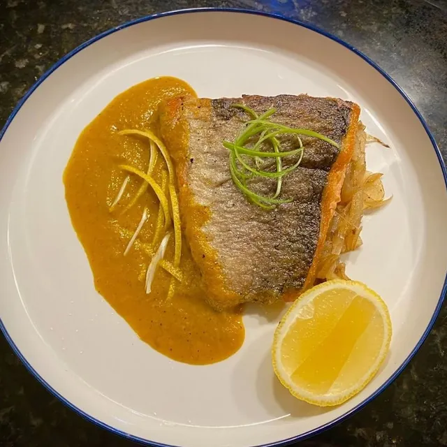 A seared salmon fillet with crisp skin, julienned scallion, an orange sauce and a lemon wedge on a blue-rimmed plate.