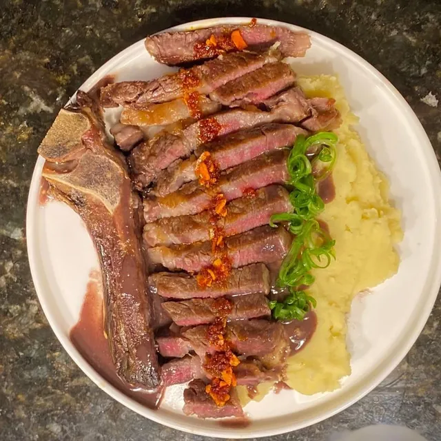 A sliced bone-in steak, pink inside, streaked with chili oil, with mashed potato and herbs on a white plate.