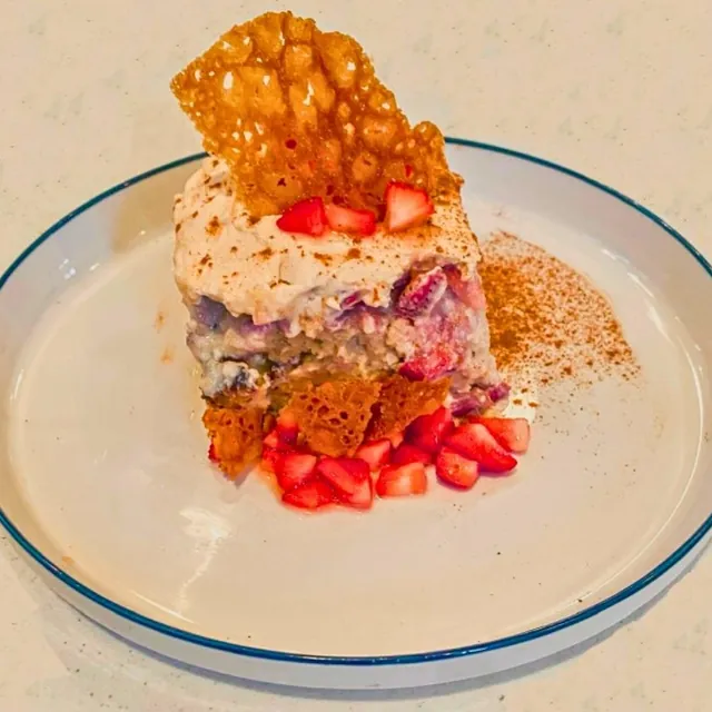 A square of layered cake under a lacy tuile, on chopped strawberries, with a dusting of red powder, on a blue-rimmed plate.