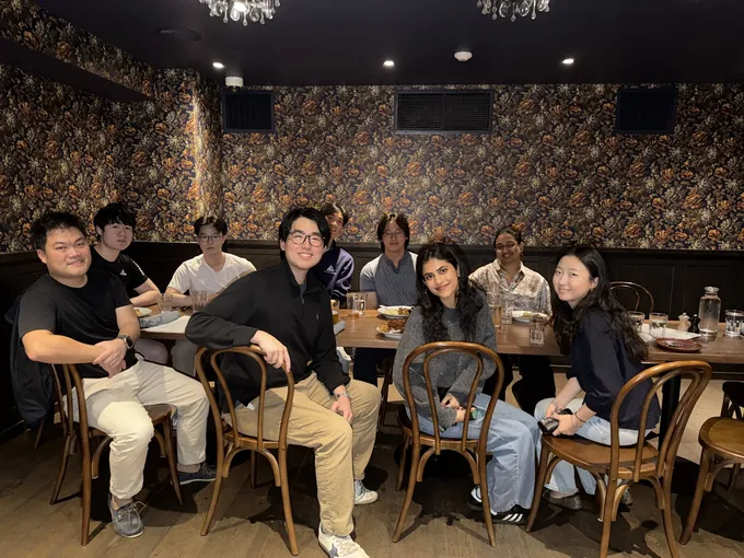 Dinner II: about a dozen guests around a long table in a restaurant with dark floral wallpaper, turned toward the camera.
