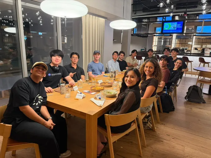 Dinner III: guests seated along a long wooden table in a bright room with tall windows, smiling at the camera.